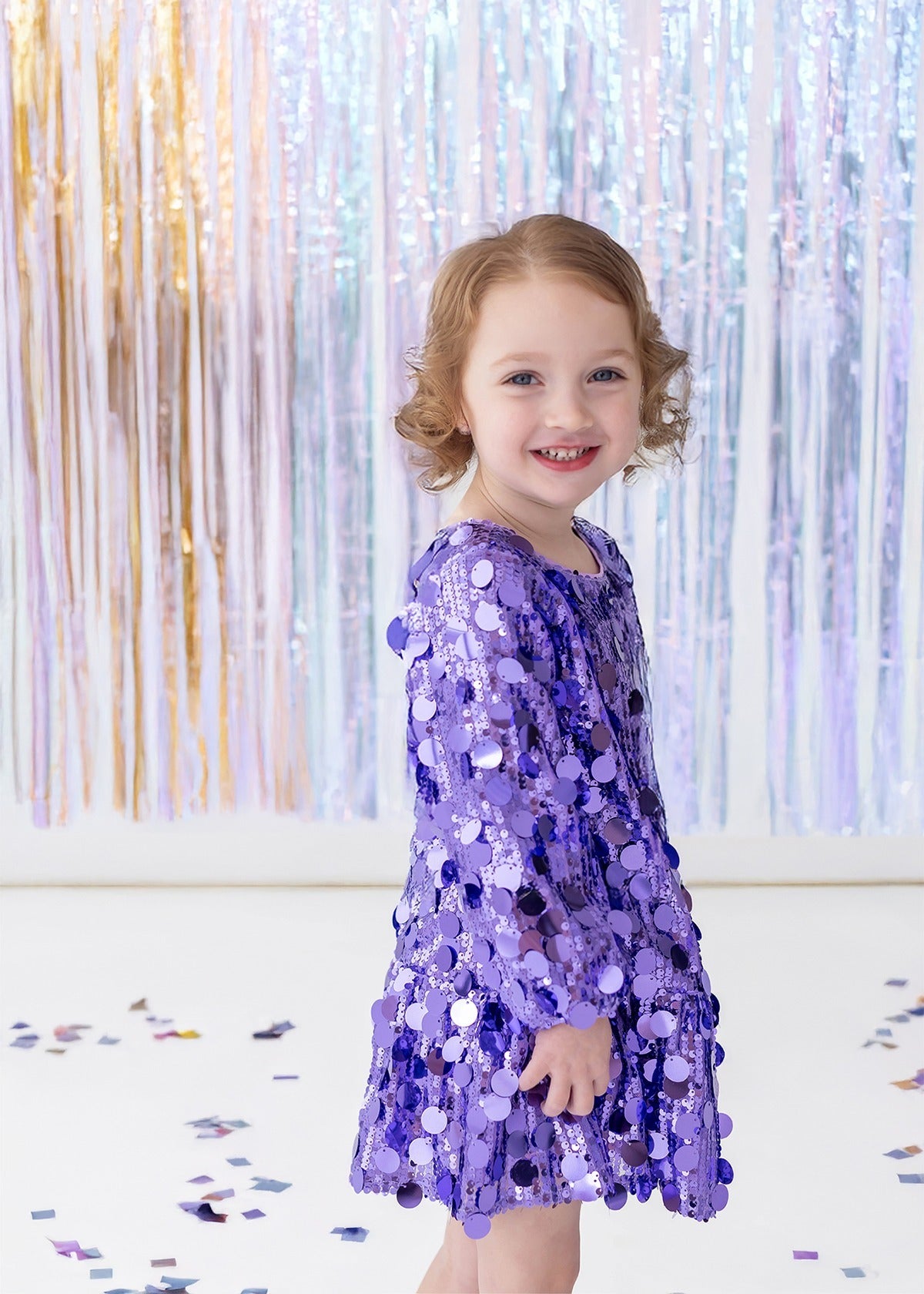 A young child wearing the Isobella and Chloe “Shake it Off” Bishop Sleeve Sequin Ruffle Hem Mini Dress smiles while standing in front of shiny pastel streamers, with confetti scattered on the white floor.