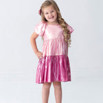 A young girl with long blonde hair, dressed in the Isobella and Chloe Berry Blush Short Sleeve Color Block Tiered Puff Sleeve Dress and a matching bow, stands smiling with her hands on her hips. She completes her look with sparkly shoes, posing against a plain white background—perfect for capturing cheerful holiday photos.