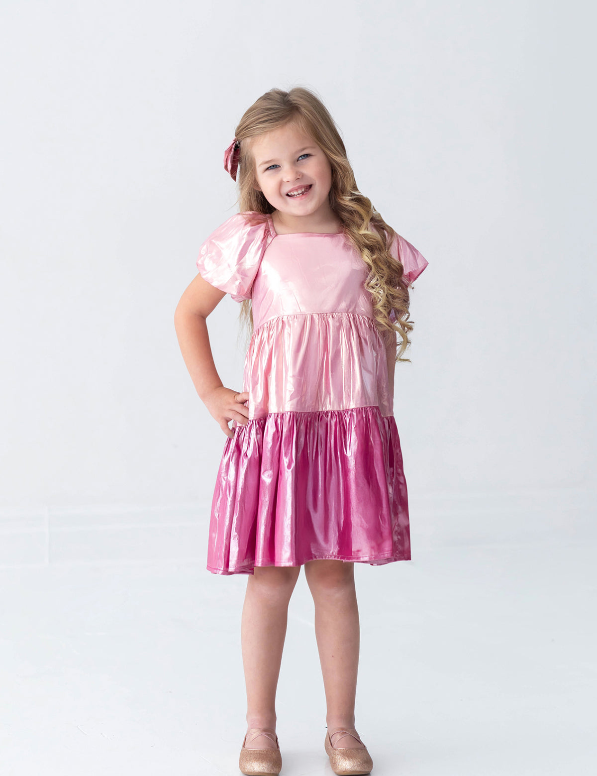 A young girl with long blonde hair, dressed in the Isobella and Chloe Berry Blush Short Sleeve Color Block Tiered Puff Sleeve Dress and a matching bow, stands smiling with her hands on her hips. She completes her look with sparkly shoes, posing against a plain white background—perfect for capturing cheerful holiday photos.