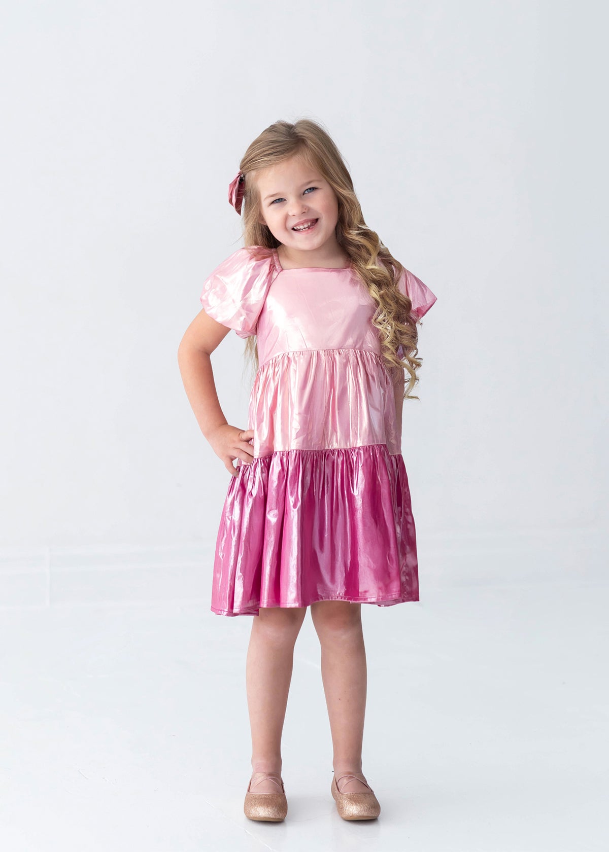 A young girl with long blonde hair, dressed in the Isobella and Chloe Berry Blush Short Sleeve Color Block Tiered Puff Sleeve Dress and a matching bow, stands smiling with her hands on her hips. She completes her look with sparkly shoes, posing against a plain white background—perfect for capturing cheerful holiday photos.