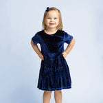 A young girl with light brown hair stands smiling, dressed in the Isobella and Chloe Velvet Dreams Short Sleeve Polka Dot Tiered Skirt Dress—a perfect special occasion outfit—paired with gray shoes and a matching blue bow in her hair, set against a light blue background.