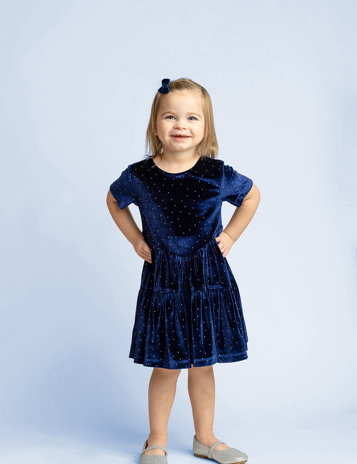 A young girl with light brown hair stands smiling, dressed in the Isobella and Chloe Velvet Dreams Short Sleeve Polka Dot Tiered Skirt Dress—a perfect special occasion outfit—paired with gray shoes and a matching blue bow in her hair, set against a light blue background.