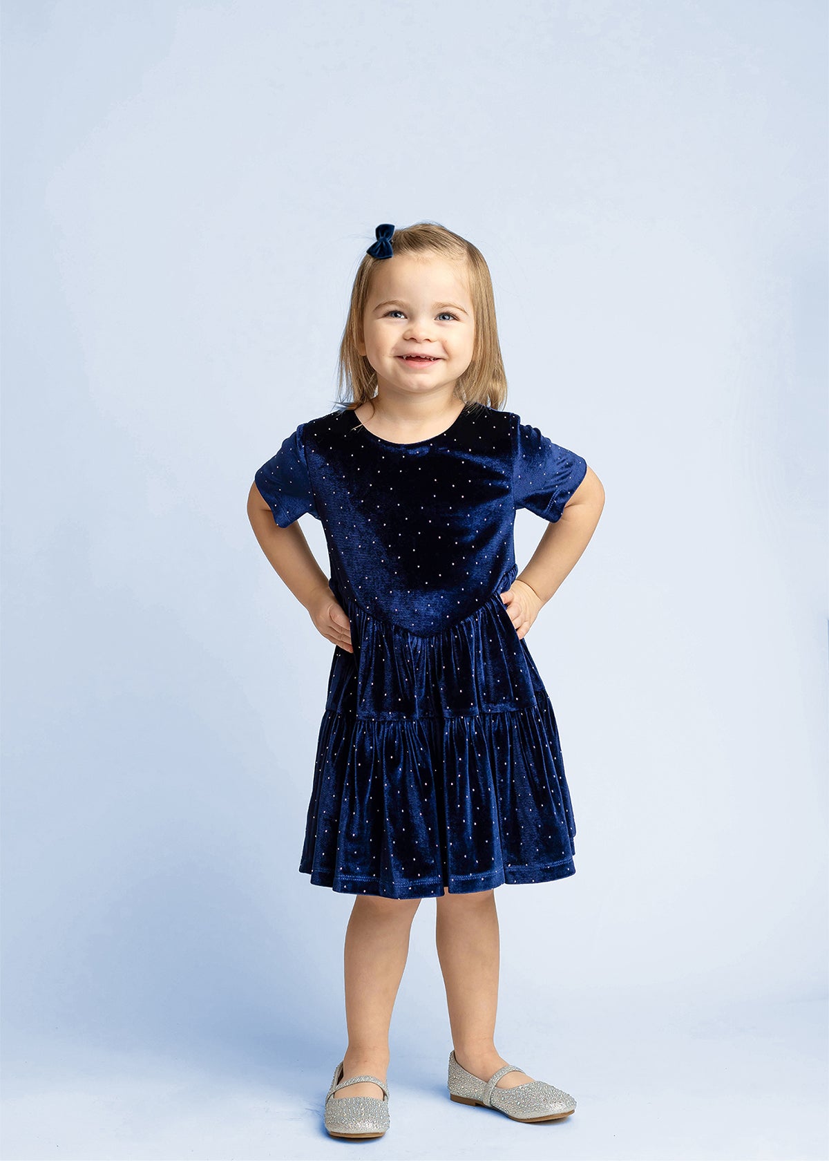 A young girl with light brown hair stands smiling, dressed in the Isobella and Chloe Velvet Dreams Short Sleeve Polka Dot Tiered Skirt Dress—a perfect special occasion outfit—paired with gray shoes and a matching blue bow in her hair, set against a light blue background.
