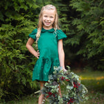 A smiling young girl stands on the grass, wearing the Isobella and Chloe Holly and Pearls Short Sleeve Solid Pearl Embellished Drop Waist Dress, holding a festive wreath adorned with greenery and red berries, with lush green trees in the background.
