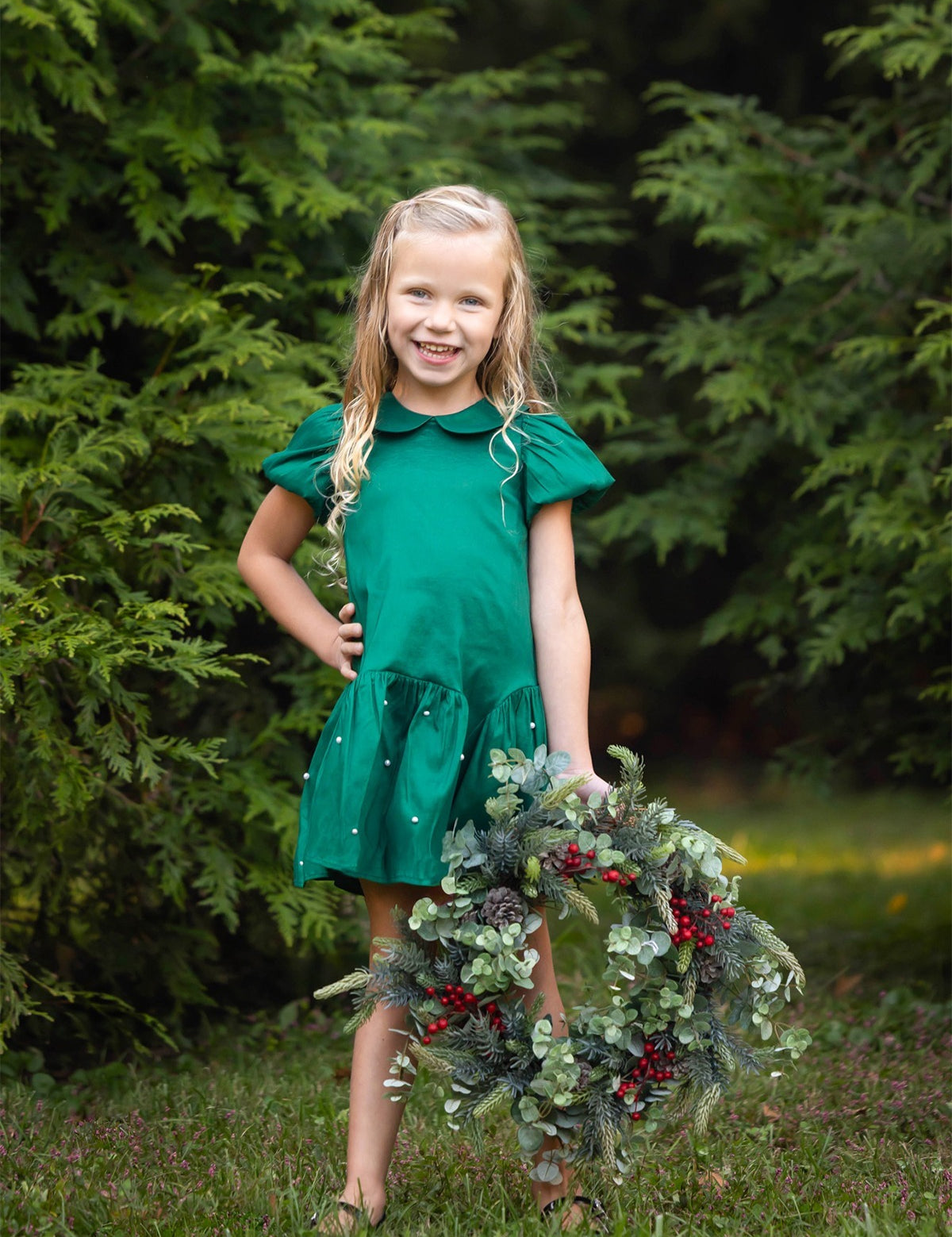 A smiling young girl stands on the grass, wearing the Isobella and Chloe Holly and Pearls Short Sleeve Solid Pearl Embellished Drop Waist Dress, holding a festive wreath adorned with greenery and red berries, with lush green trees in the background.