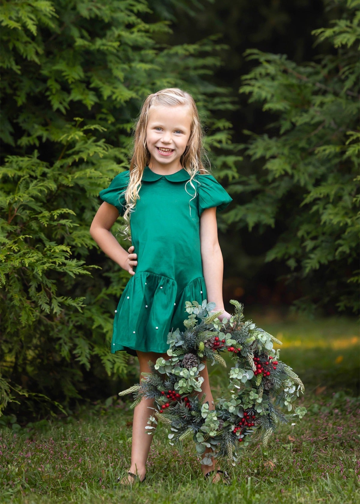 A smiling young girl stands on the grass, wearing the Isobella and Chloe Holly and Pearls Short Sleeve Solid Pearl Embellished Drop Waist Dress, holding a festive wreath adorned with greenery and red berries, with lush green trees in the background.