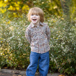 A young child with light brown hair, wearing the Beckett and Bear Nicholas Long Sleeve Plaid Chest Pocket Button Front Shirt and blue jeans, stands outdoors on a path, smiling with hands behind their back. Bushes and greenery fill the background.