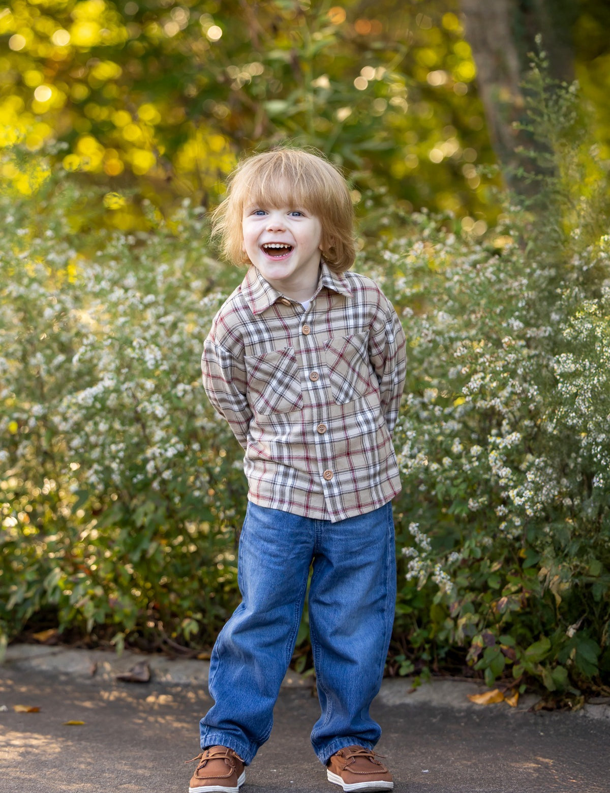 A young child with light brown hair, wearing the Beckett and Bear Nicholas Long Sleeve Plaid Chest Pocket Button Front Shirt and blue jeans, stands outdoors on a path, smiling with hands behind their back. Bushes and greenery fill the background.