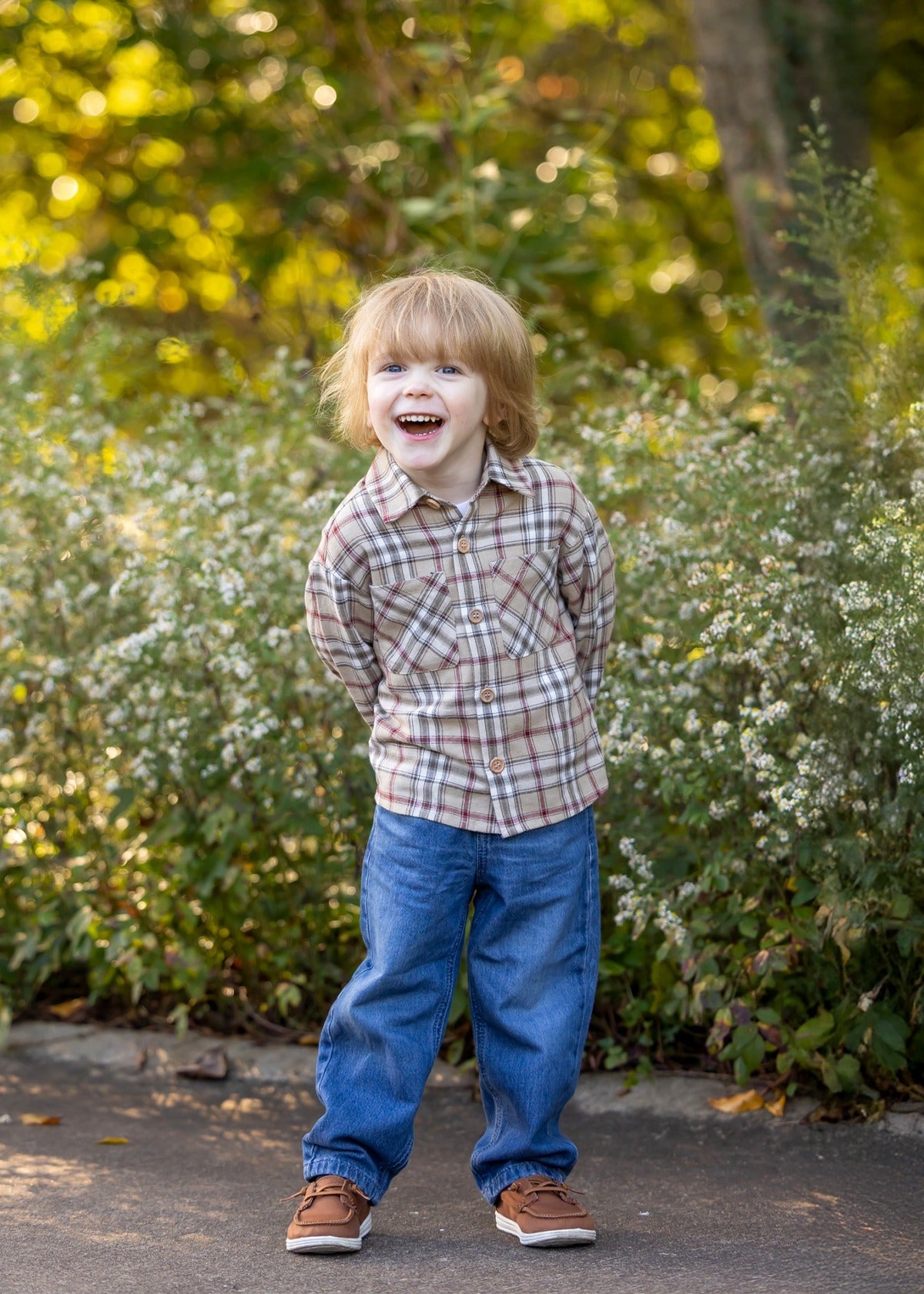 A young child with light brown hair, wearing the Beckett and Bear Nicholas Long Sleeve Plaid Chest Pocket Button Front Shirt and blue jeans, stands outdoors on a path, smiling with hands behind their back. Bushes and greenery fill the background.