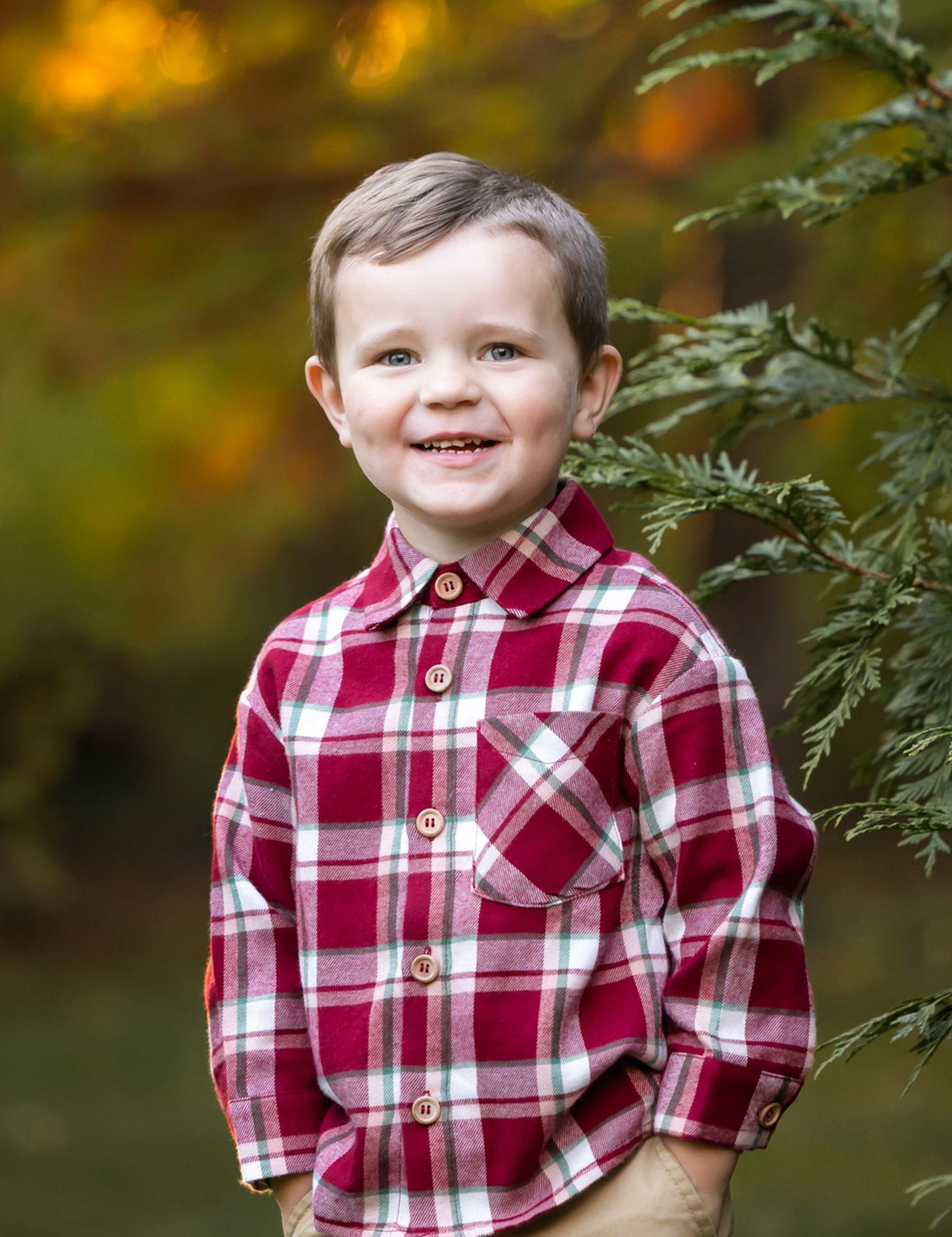 A smiling young boy with short brown hair wears the Beckett and Bear Joseph Long Sleeve Plaid Flannel Chest Pocket Shirt and khaki pants, standing outdoors by a green tree branch with a softly blurred autumn background.