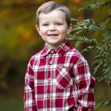 A smiling young boy with short brown hair wears the Beckett and Bear Joseph Long Sleeve Plaid Flannel Chest Pocket Shirt and khaki pants, standing outdoors by a green tree branch with a softly blurred autumn background.