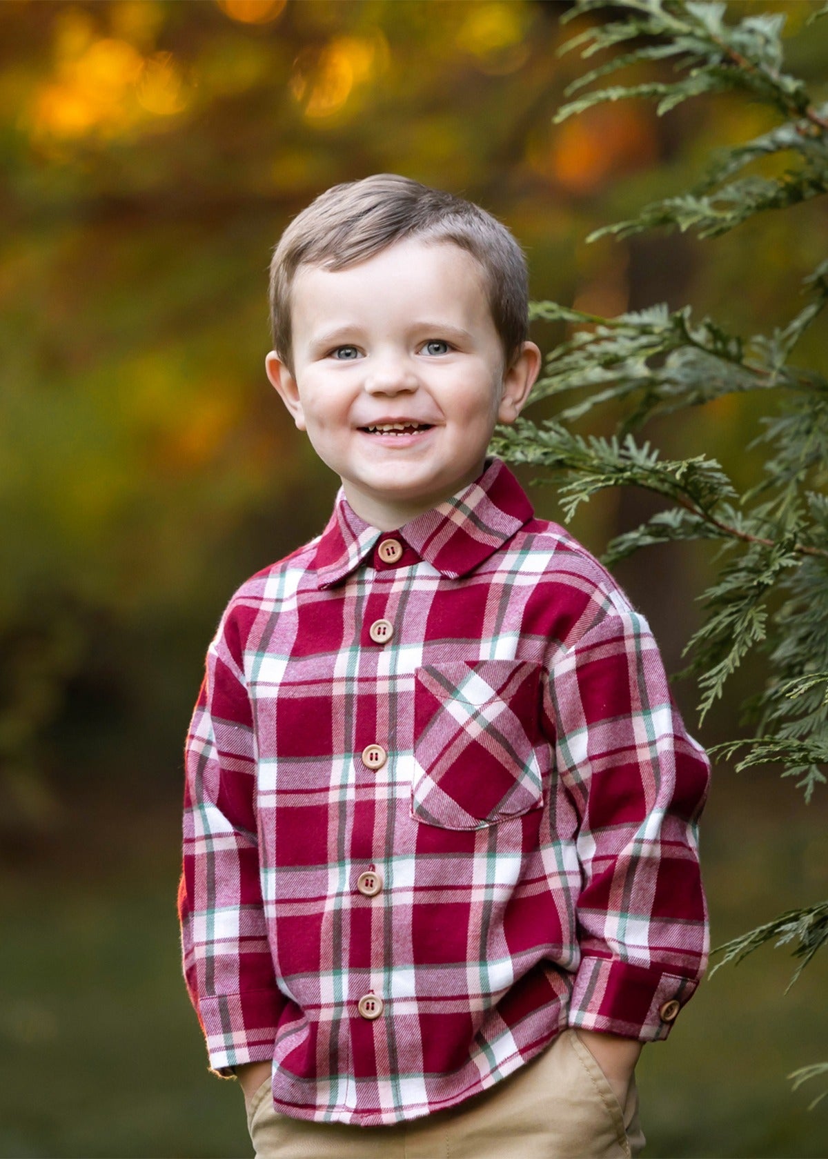 A smiling young boy with short brown hair wears the Beckett and Bear Joseph Long Sleeve Plaid Flannel Chest Pocket Shirt and khaki pants, standing outdoors by a green tree branch with a softly blurred autumn background.