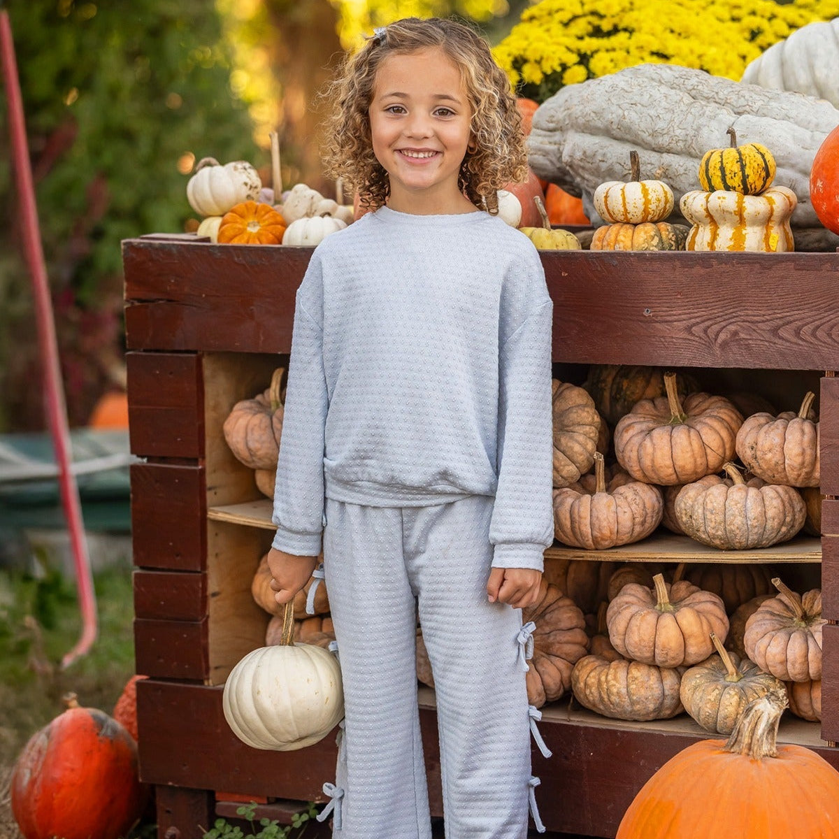A smiling child with curly hair stands in front of a wooden display filled with pumpkins and gourds, holding a white pumpkin in one hand. The playful Eva Long Sleeve Solid Bow Tie Top and Pant Set by Mabel and Honey, crafted from a dot knit cotton blend fabric and featuring wide leg pants, adds comfort and charm to her look. Yellow flowers and greenery brighten the background.