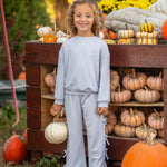 A smiling child with curly hair stands in front of a wooden display filled with pumpkins and gourds, holding a white pumpkin in one hand. The playful Eva Long Sleeve Solid Bow Tie Top and Pant Set by Mabel and Honey, crafted from a dot knit cotton blend fabric and featuring wide leg pants, adds comfort and charm to her look. Yellow flowers and greenery brighten the background.