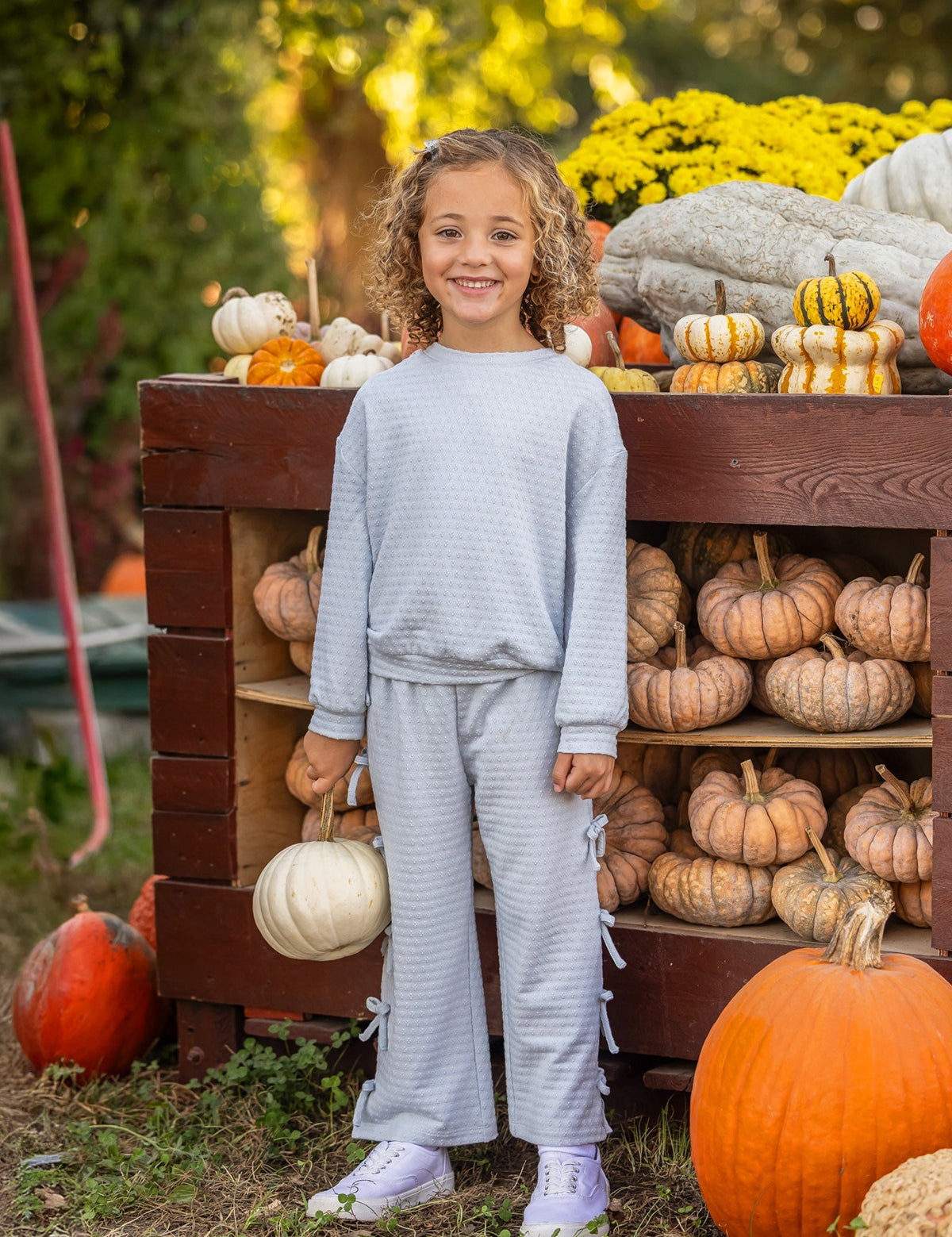 A smiling child with curly hair stands in front of a wooden display filled with pumpkins and gourds, holding a white pumpkin in one hand. The playful Eva Long Sleeve Solid Bow Tie Top and Pant Set by Mabel and Honey, crafted from a dot knit cotton blend fabric and featuring wide leg pants, adds comfort and charm to her look. Yellow flowers and greenery brighten the background.