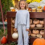 A smiling child with curly hair stands in front of a wooden display filled with pumpkins and gourds, holding a white pumpkin in one hand. The playful Eva Long Sleeve Solid Bow Tie Top and Pant Set by Mabel and Honey, crafted from a dot knit cotton blend fabric and featuring wide leg pants, adds comfort and charm to her look. Yellow flowers and greenery brighten the background.
