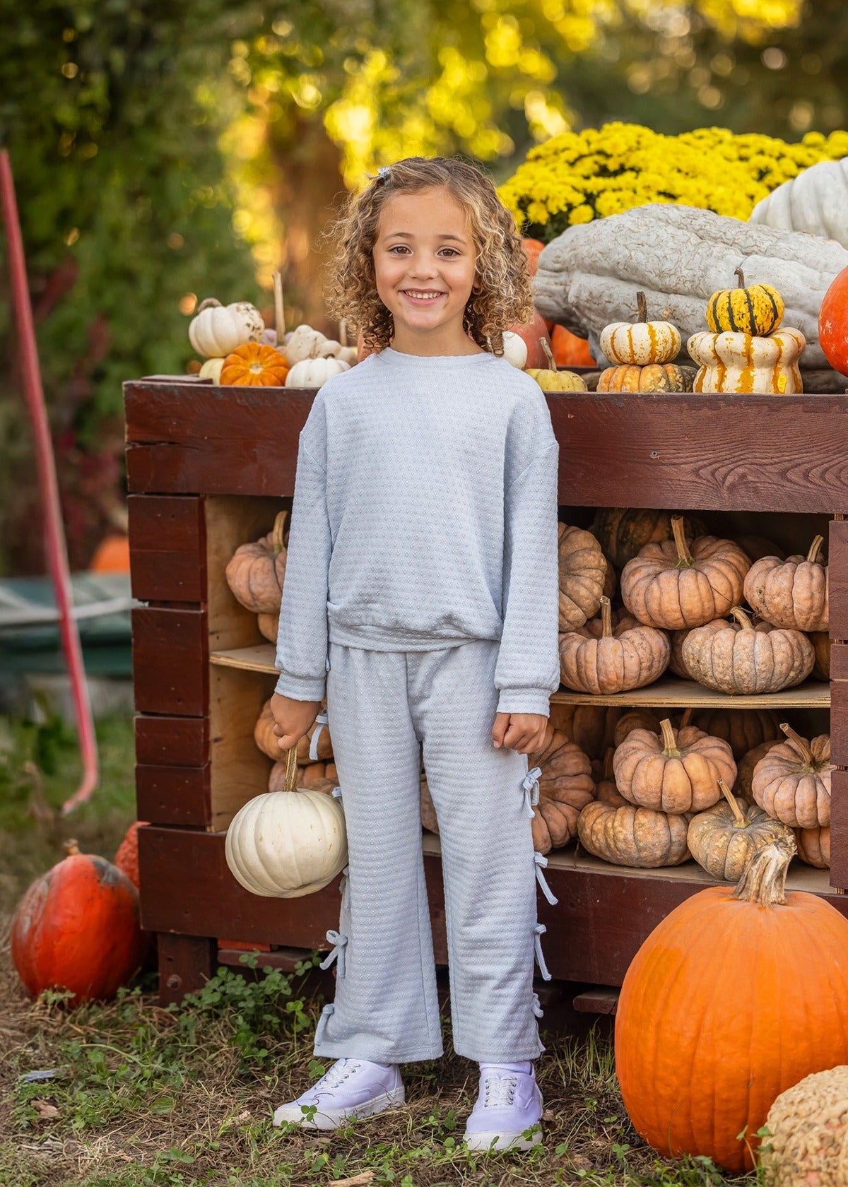 A smiling child with curly hair stands in front of a wooden display filled with pumpkins and gourds, holding a white pumpkin in one hand. The playful Eva Long Sleeve Solid Bow Tie Top and Pant Set by Mabel and Honey, crafted from a dot knit cotton blend fabric and featuring wide leg pants, adds comfort and charm to her look. Yellow flowers and greenery brighten the background.