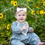 A smiling baby wearing the Dolly Long Sleeve Floral Lace Heart Bubble Romper by Mabel and Honey, featuring a dusty blue floral print in lightweight cotton with a delicate lace heart applique, sits outdoors among yellow flowers and greenery, topped with a white bow headband.