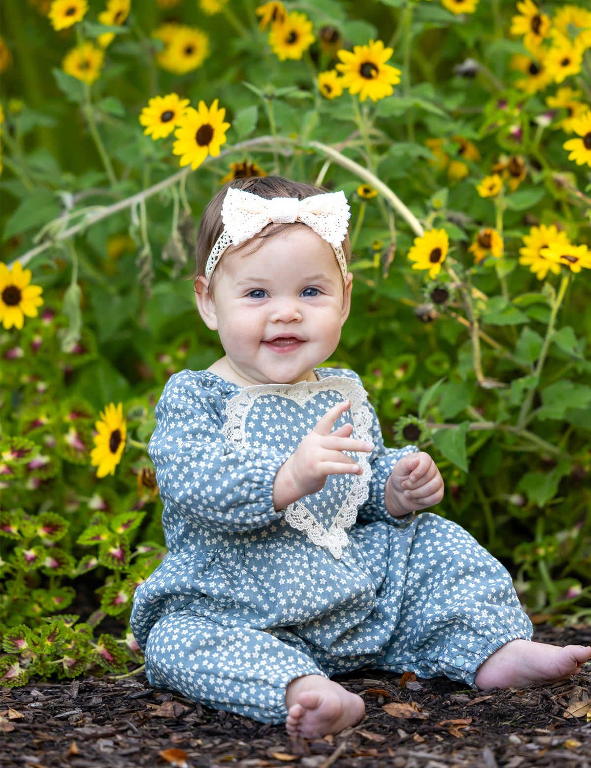 A smiling baby wearing the Dolly Long Sleeve Floral Lace Heart Bubble Romper by Mabel and Honey, featuring a dusty blue floral print in lightweight cotton with a delicate lace heart applique, sits outdoors among yellow flowers and greenery, topped with a white bow headband.