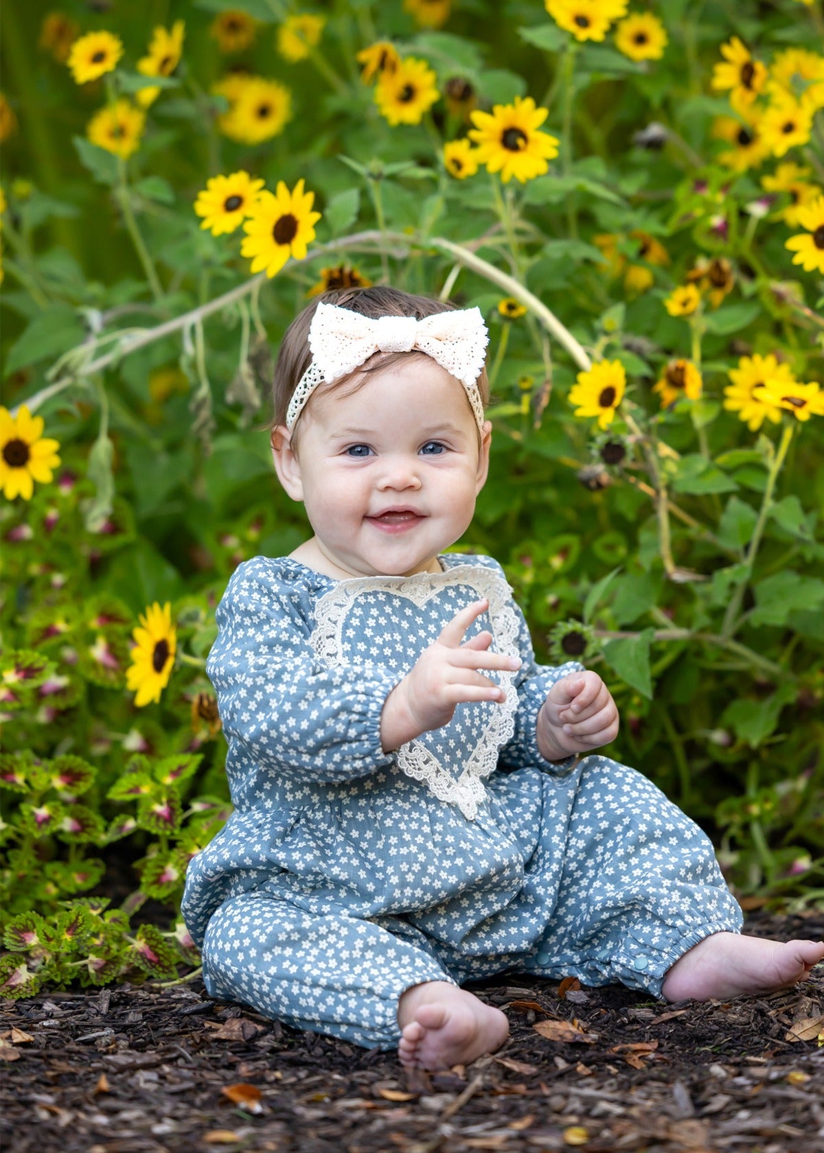 A smiling baby wearing the Dolly Long Sleeve Floral Lace Heart Bubble Romper by Mabel and Honey, featuring a dusty blue floral print in lightweight cotton with a delicate lace heart applique, sits outdoors among yellow flowers and greenery, topped with a white bow headband.