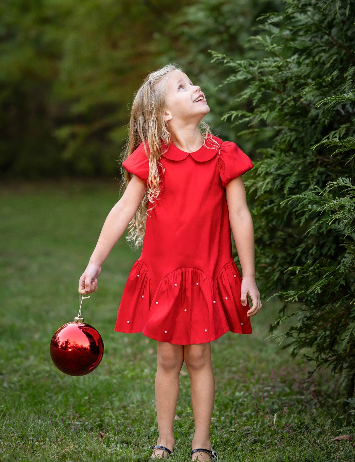A young girl wearing the Isobella and Chloe Crimson Glow Short Sleeve Peter Pan Collar Pearl Embellished Dropwaist Dress stands on the grass holding a large red ornament, gazing upward amid lush greenery—her festive, pearl-accented crimson dress perfect for holiday parties.