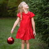 A young girl wearing the Isobella and Chloe Crimson Glow Short Sleeve Peter Pan Collar Pearl Embellished Dropwaist Dress stands on the grass holding a large red ornament, gazing upward amid lush greenery—her festive, pearl-accented crimson dress perfect for holiday parties.