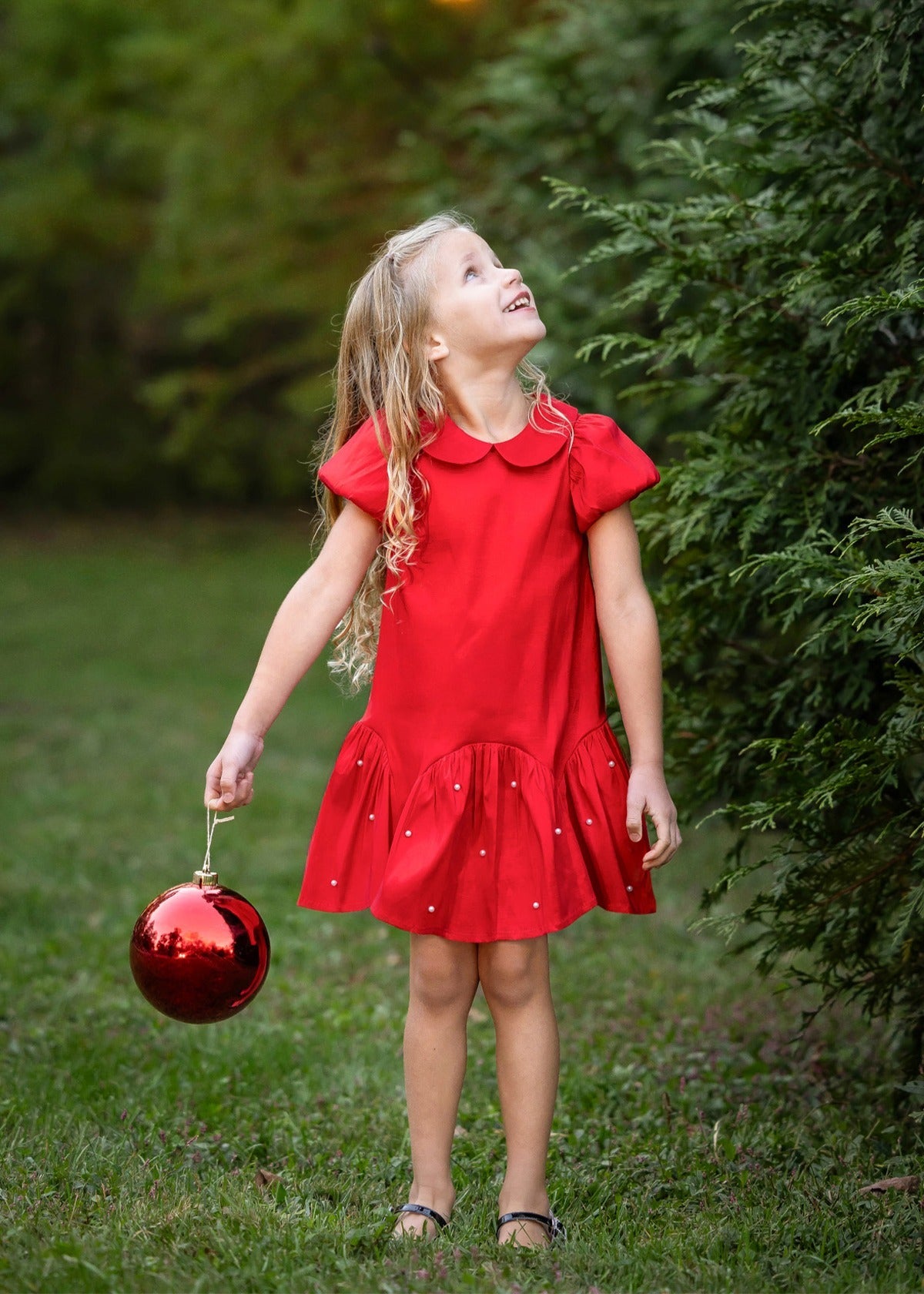 A young girl wearing the Isobella and Chloe Crimson Glow Short Sleeve Peter Pan Collar Pearl Embellished Dropwaist Dress stands on the grass holding a large red ornament, gazing upward amid lush greenery—her festive, pearl-accented crimson dress perfect for holiday parties.
