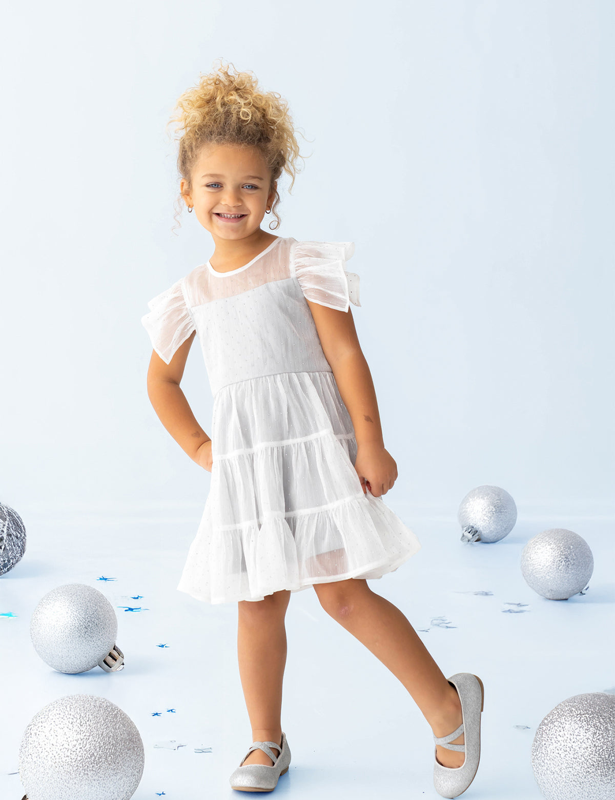 A young child wearing the Isobella and Chloe Shimmer Shine Flutter Sleeve Polka Dot Tiered Dress and gray shoes stands smiling on a light blue floor, surrounded by large silver and blue decorative balls.