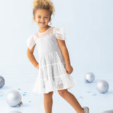 A young child wearing the Isobella and Chloe Shimmer Shine Flutter Sleeve Polka Dot Tiered Dress and gray shoes stands smiling on a light blue floor, surrounded by large silver and blue decorative balls.