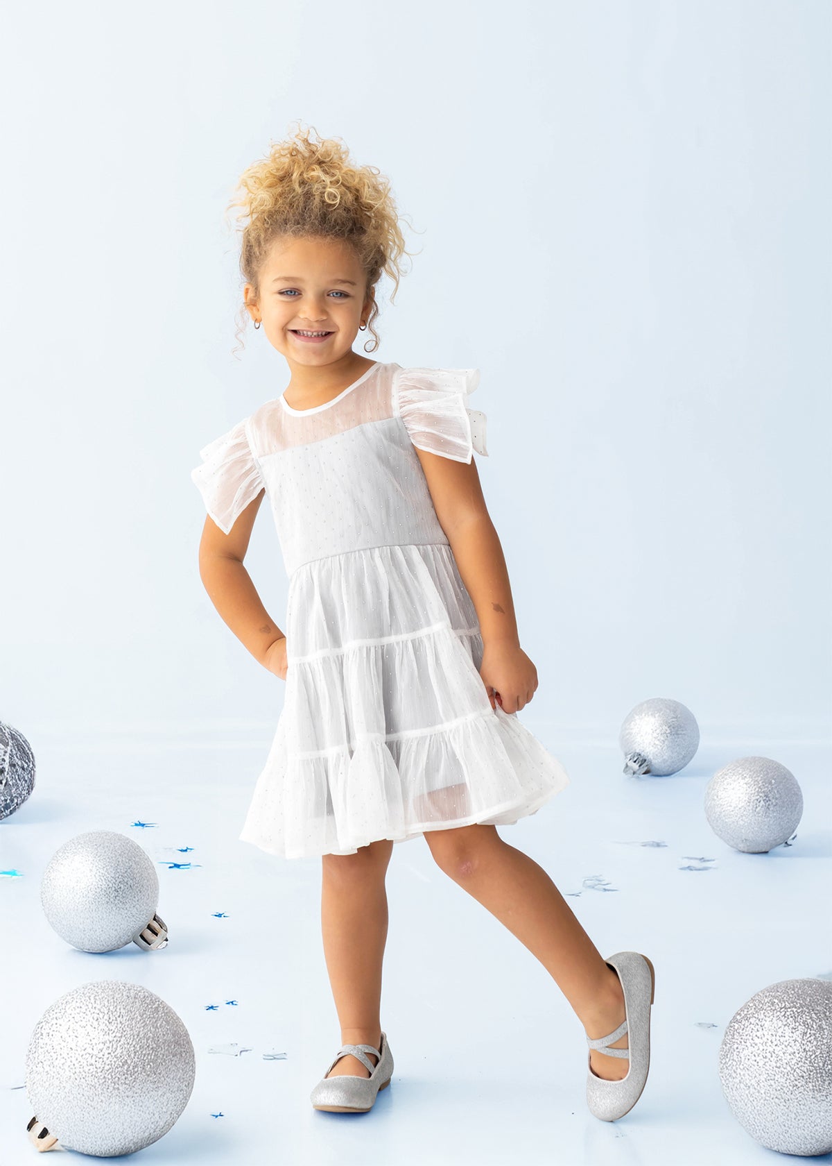 A young child wearing the Isobella and Chloe Shimmer Shine Flutter Sleeve Polka Dot Tiered Dress and gray shoes stands smiling on a light blue floor, surrounded by large silver and blue decorative balls.
