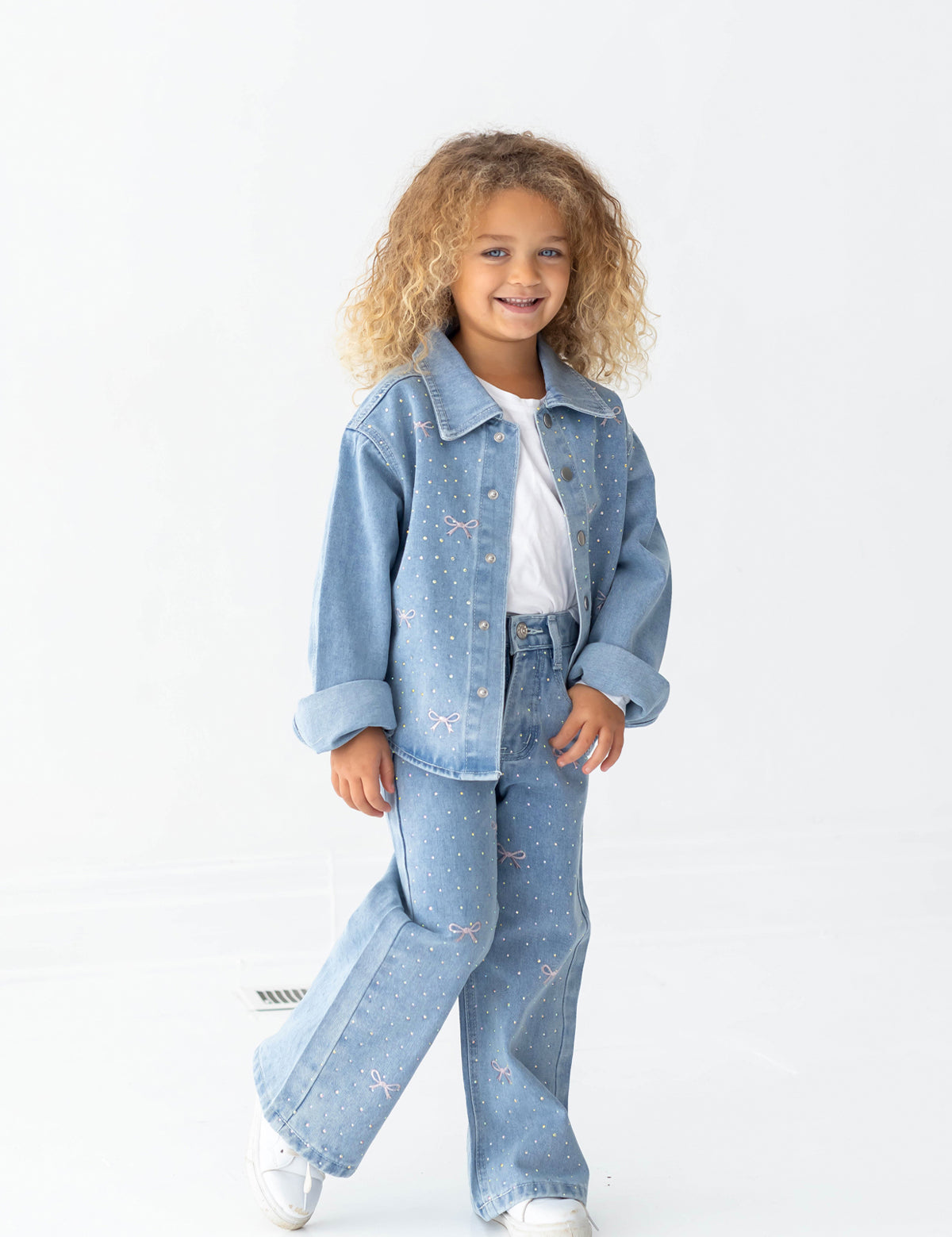 A young child with curly blonde hair smiles while wearing the Mabel and Honey Long Sleeve Embroidered Bow Rhinestone Dot Denim Jacket, paired with matching jeans, a white t-shirt, and white sneakers, standing against a plain white background.