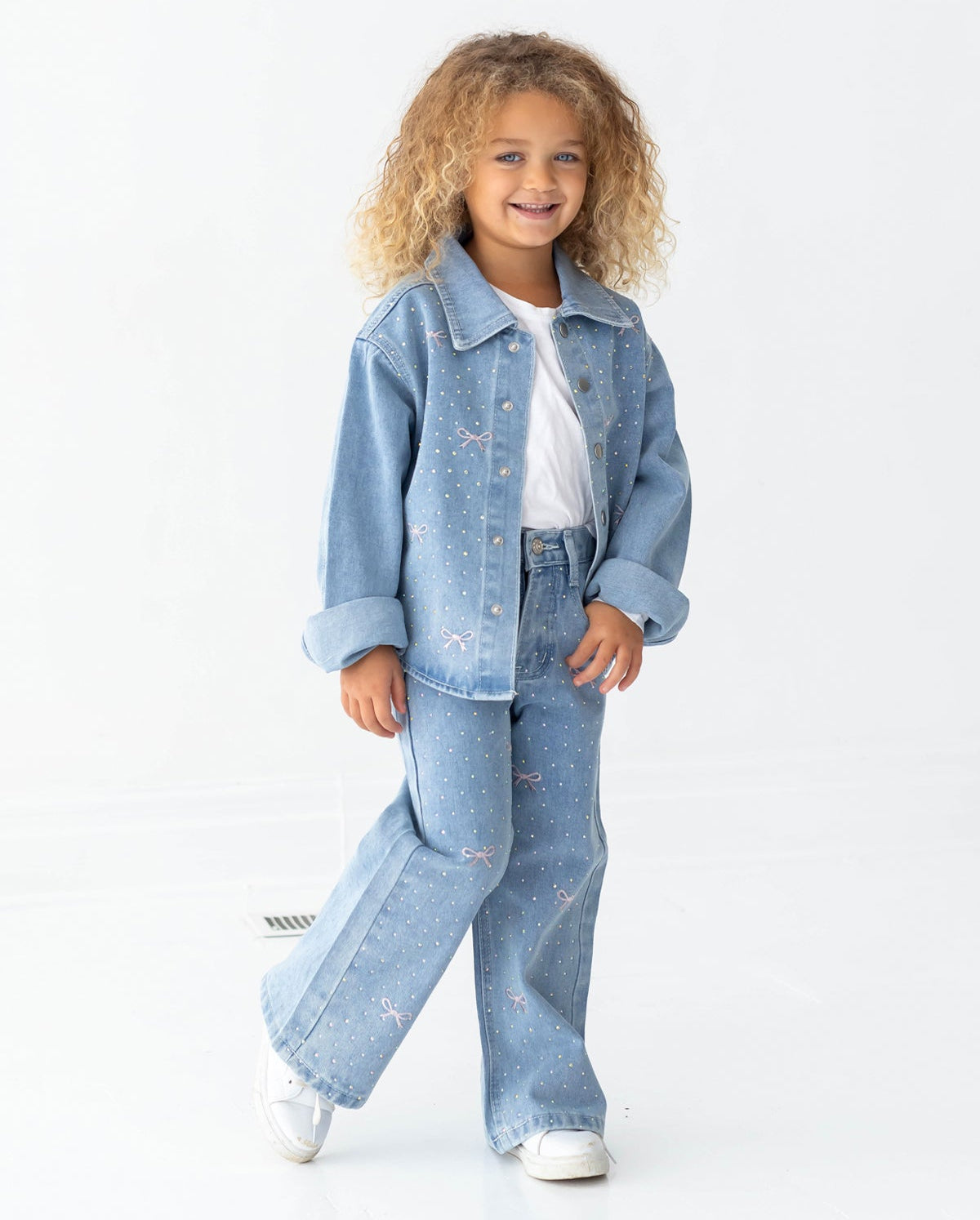 A young child with curly hair smiles, dressed in a light blue denim jacket and Mabel and Honey Bow Embroidered Rhinestone Wide Leg Denim Pants for toddlers and little kids, which feature small dots, pink bow embroidery, and a wide leg silhouette. The look is completed with a white shirt and white sneakers, set against a plain white background.
