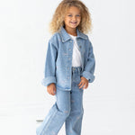 A young child with curly hair smiles while wearing the Bow Embroidered Rhinestone Wide Leg Denim Pants from Mabel and Honey, paired with a light blue denim jacket, a white t-shirt, and white sneakers. The stylish light wash pants feature a playful wide leg silhouette adorned with small colorful bow embellishments, perfectly suited for a cheerful look against a plain white background.