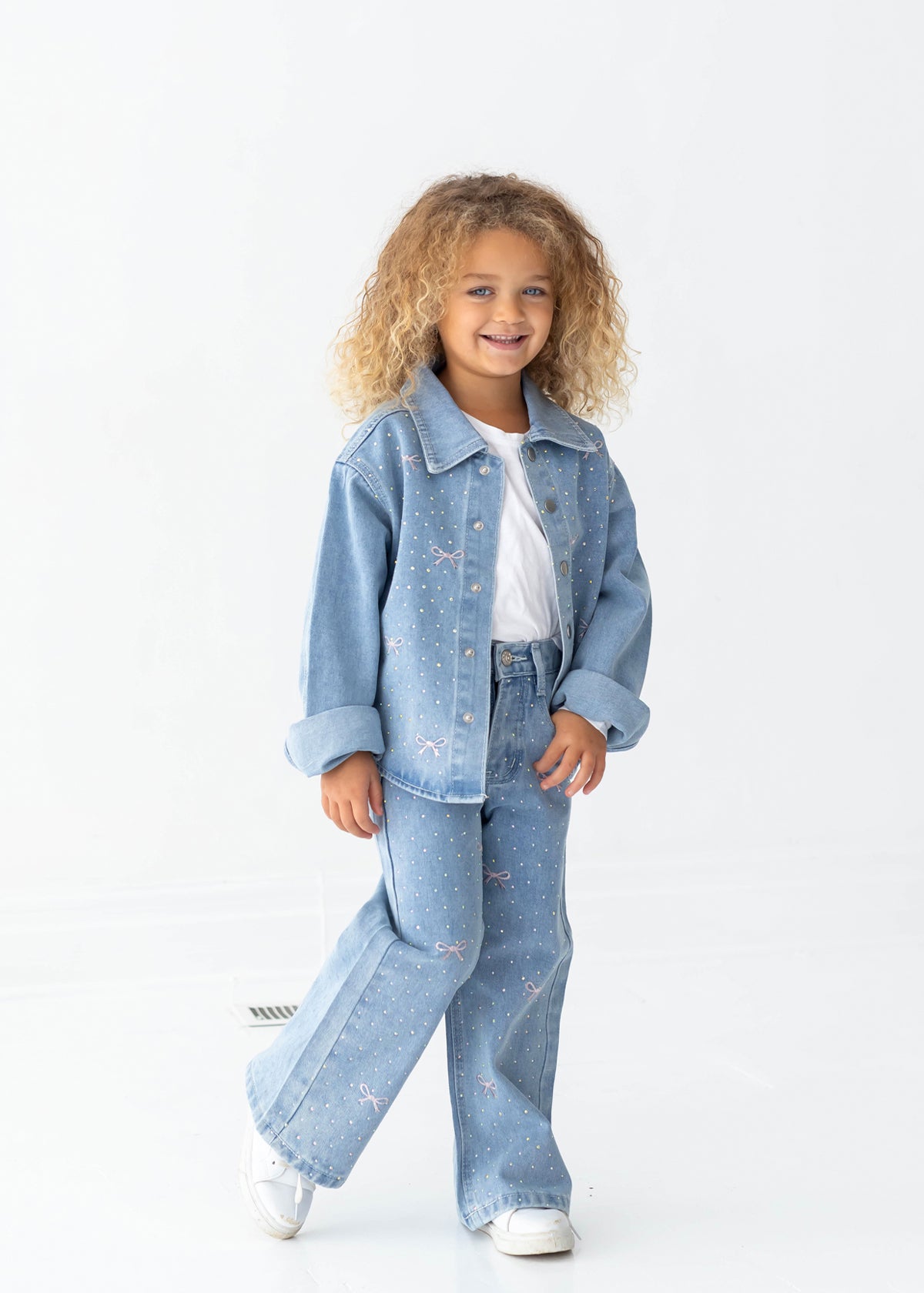 A young child with curly hair smiles while wearing the Bow Embroidered Rhinestone Wide Leg Denim Pants from Mabel and Honey, paired with a light blue denim jacket, a white t-shirt, and white sneakers. The stylish light wash pants feature a playful wide leg silhouette adorned with small colorful bow embellishments, perfectly suited for a cheerful look against a plain white background.