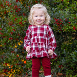 Wearing the Mabel and Honey Winterberry Long Sleeve Plaid Tiered Ruffle Top and Legging Set, a young child with blonde hair stands smiling in front of a bush with red berries, her brown boots completing this perfect girls’ holiday outfit.