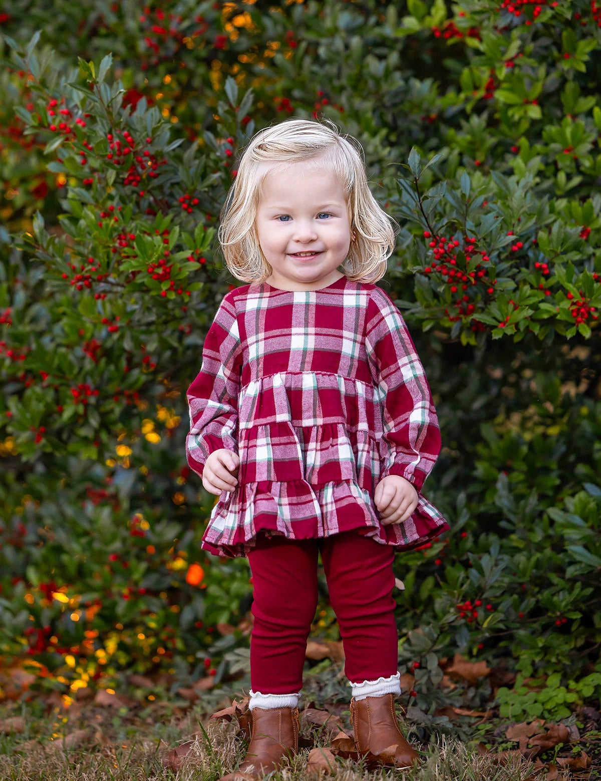 Wearing the Mabel and Honey Winterberry Long Sleeve Plaid Tiered Ruffle Top and Legging Set, a young child with blonde hair stands smiling in front of a bush with red berries, her brown boots completing this perfect girls’ holiday outfit.