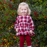 Wearing the Mabel and Honey Winterberry Long Sleeve Plaid Tiered Ruffle Top and Legging Set, a young child with blonde hair stands smiling in front of a bush with red berries, her brown boots completing this perfect girls’ holiday outfit.