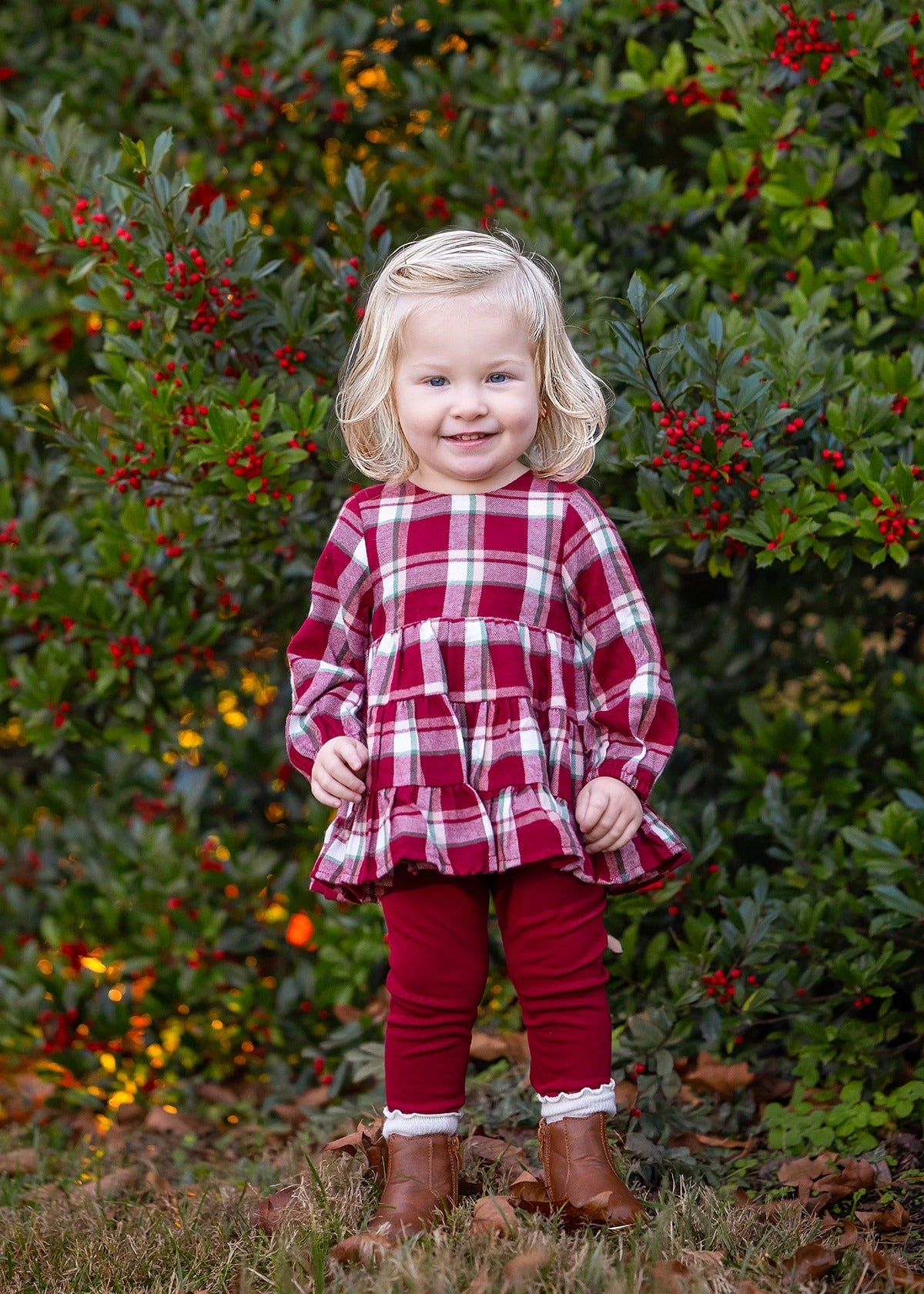 Wearing the Mabel and Honey Winterberry Long Sleeve Plaid Tiered Ruffle Top and Legging Set, a young child with blonde hair stands smiling in front of a bush with red berries, her brown boots completing this perfect girls’ holiday outfit.