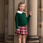 A young girl with long, wavy blonde hair stands smiling by stone columns. She wears a green sweater, a white-collared shirt, and the Mabel and Honey Winterberry Plaid Elastic Waist Brushed Flannel Raw Edge Hem Skirt, complemented by white ankle boots.