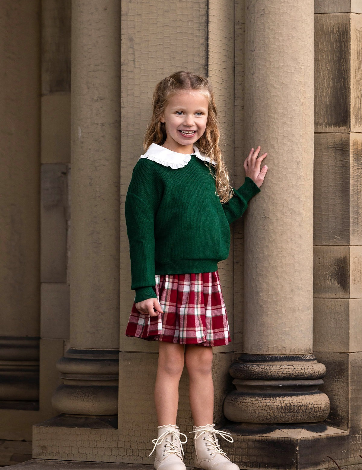 A young girl with long, wavy blonde hair stands smiling by stone columns. She wears a green sweater, a white-collared shirt, and the Mabel and Honey Winterberry Plaid Elastic Waist Brushed Flannel Raw Edge Hem Skirt, complemented by white ankle boots.