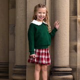 A young girl with long, wavy blonde hair stands smiling by stone columns. She wears a green sweater, a white-collared shirt, and the Mabel and Honey Winterberry Plaid Elastic Waist Brushed Flannel Raw Edge Hem Skirt, complemented by white ankle boots.