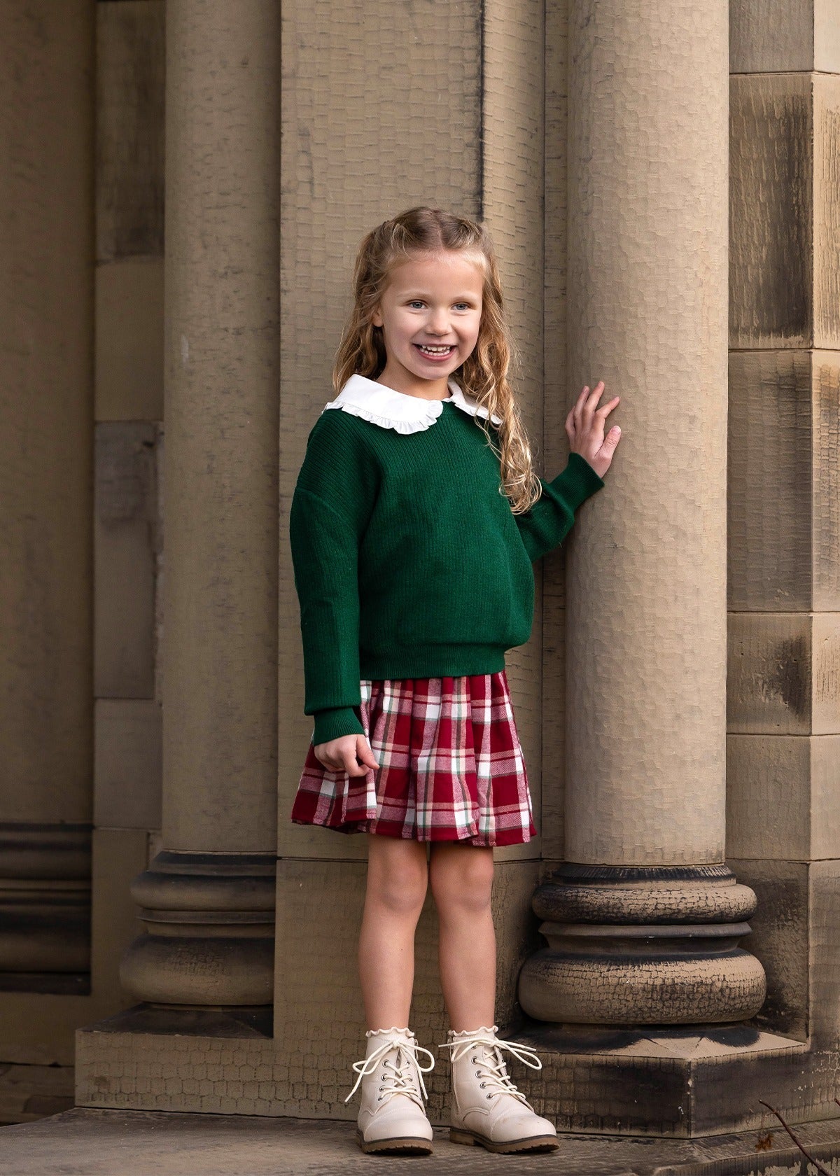 A young girl with long, wavy blonde hair stands smiling by stone columns. She wears a green sweater, a white-collared shirt, and the Mabel and Honey Winterberry Plaid Elastic Waist Brushed Flannel Raw Edge Hem Skirt, complemented by white ankle boots.