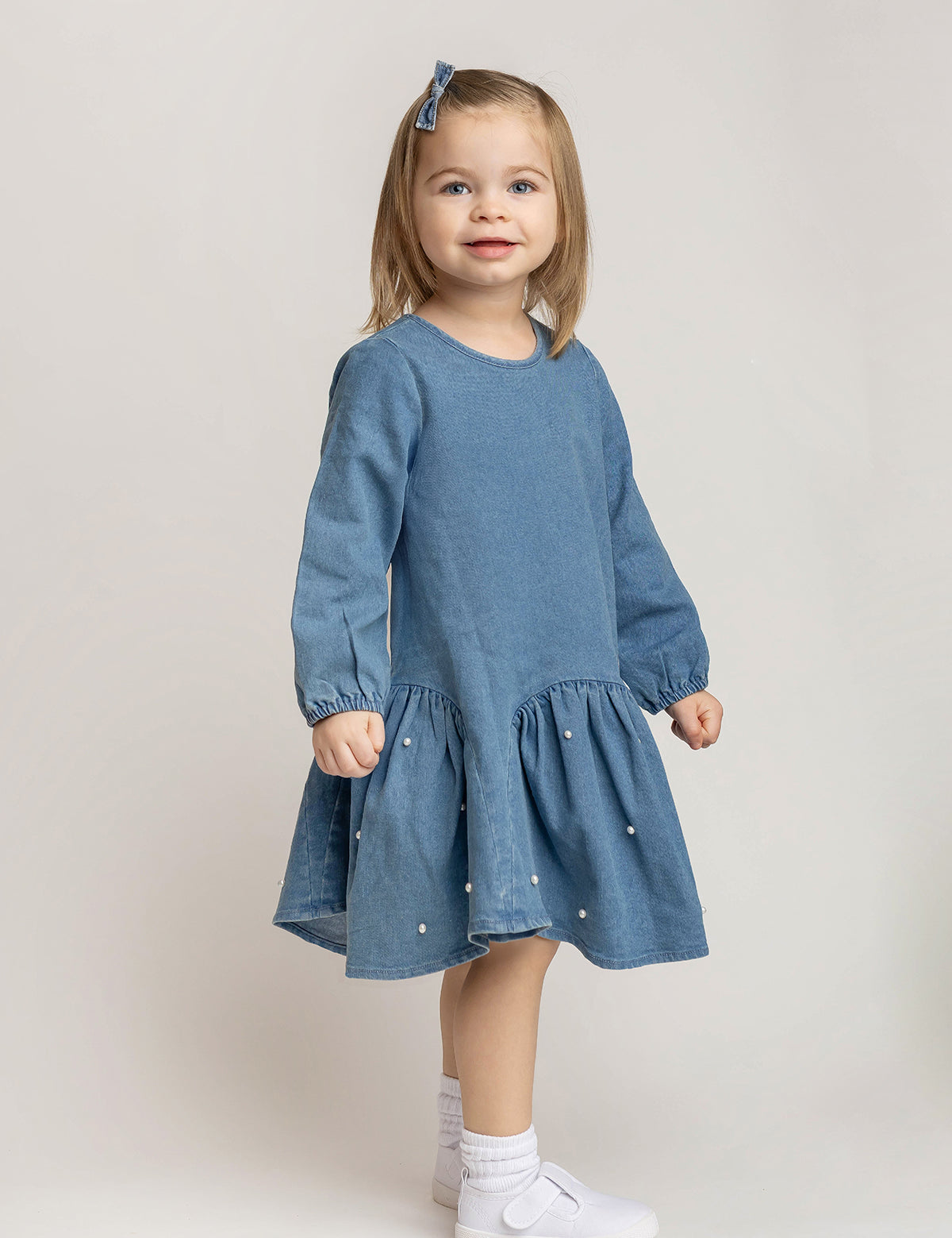 A young girl with light brown hair wears the Mabel and Honey Denim Doll Long Sleeve Solid Pearl Embellished Dropwaist Dress, paired with white socks, white shoes, and a coordinating blue hair bow, as she stands smiling against a light background.