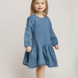 A young girl with light brown hair wears the Mabel and Honey Denim Doll Long Sleeve Solid Pearl Embellished Dropwaist Dress, paired with white socks, white shoes, and a coordinating blue hair bow, as she stands smiling against a light background.