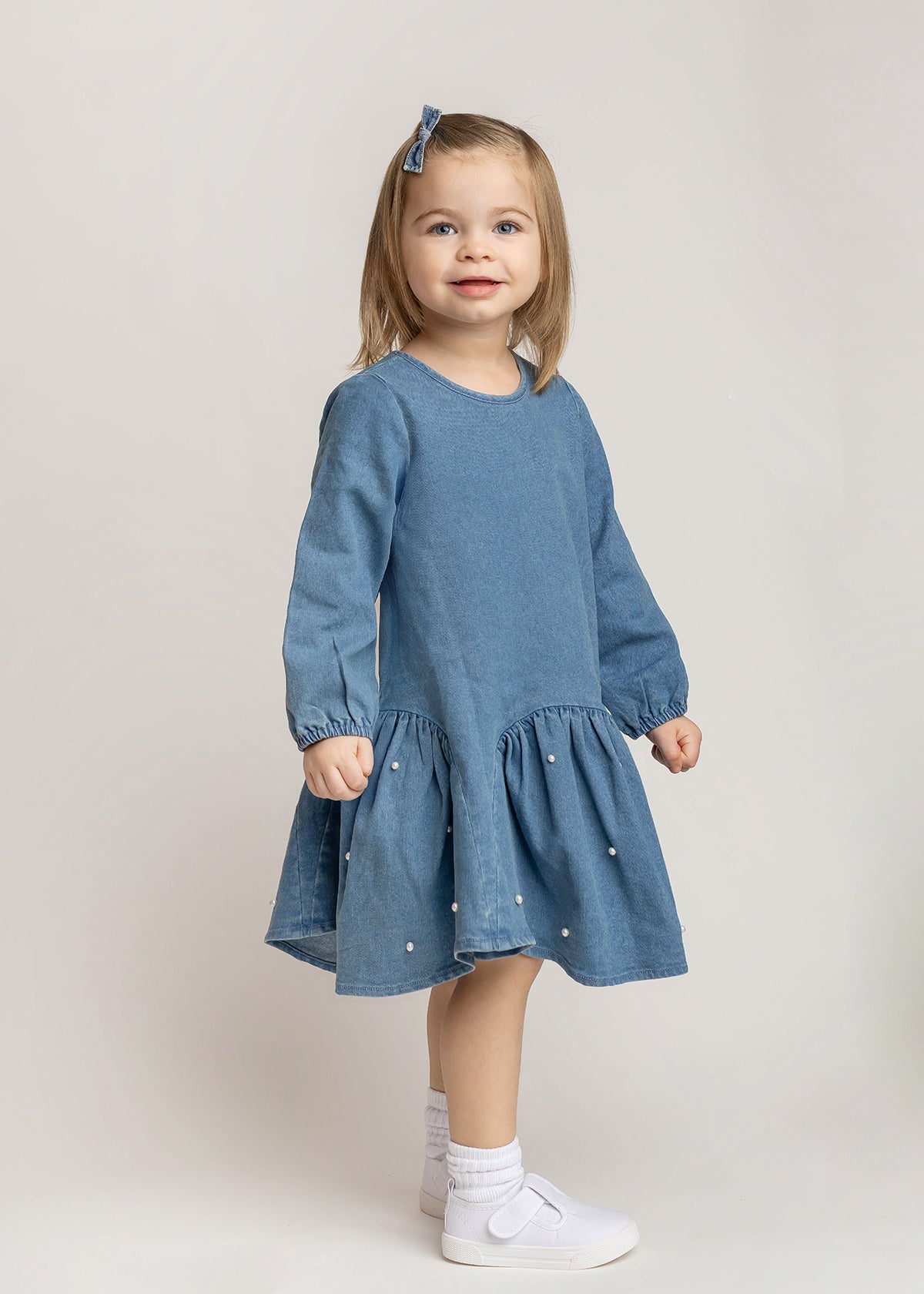 A young girl with light brown hair wears the Mabel and Honey Denim Doll Long Sleeve Solid Pearl Embellished Dropwaist Dress, paired with white socks, white shoes, and a coordinating blue hair bow, as she stands smiling against a light background.