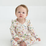 A baby with light hair sits barefoot on a white floor, wearing cream-colored Cactus Kisses Western Print Elastic Waist Leggings by Mabel and Honey, adorned with colorful animals and playful western-inspired patterns, gazing slightly upward with a curious expression.