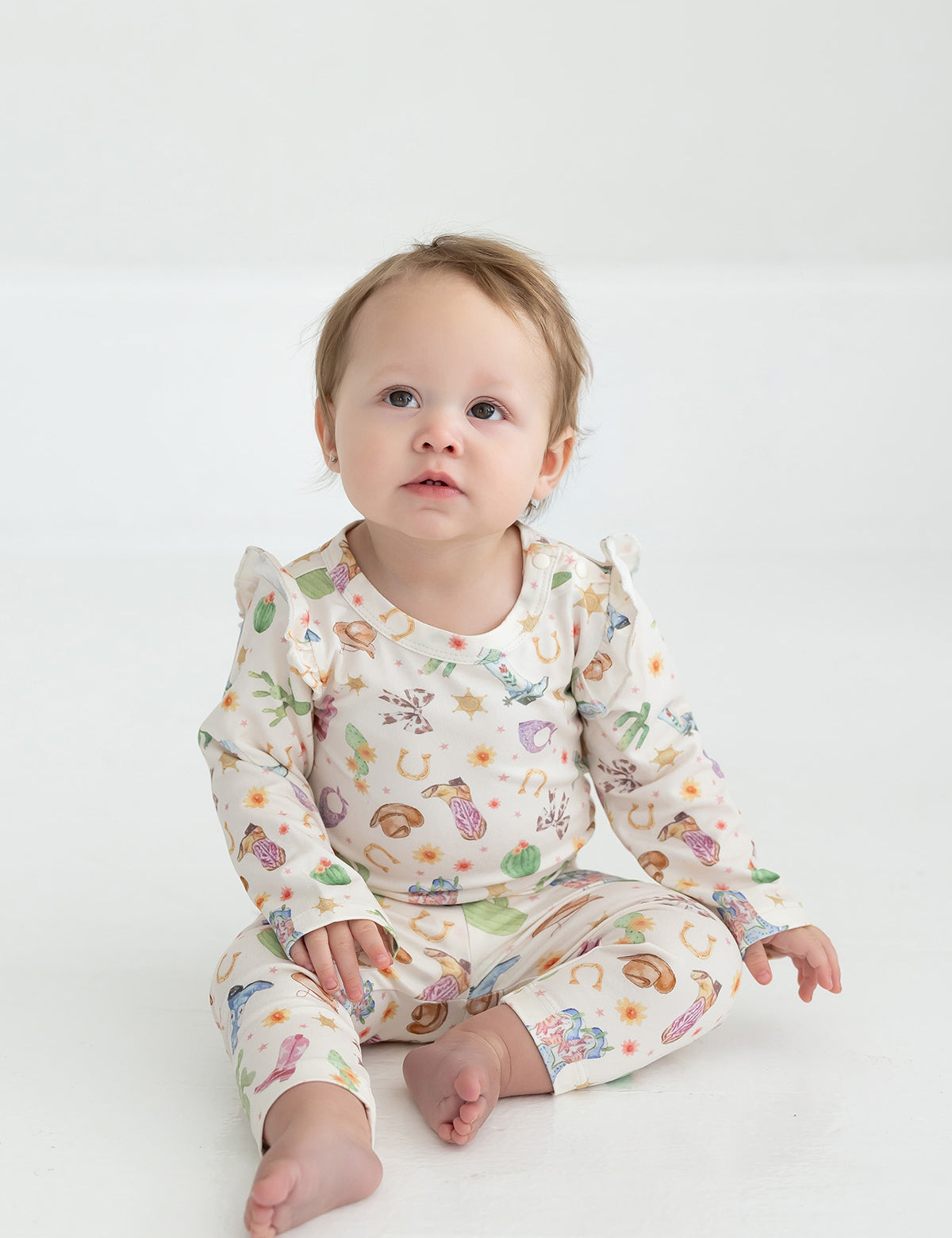 A baby with light hair sits barefoot on a white floor, wearing cream-colored Cactus Kisses Western Print Elastic Waist Leggings by Mabel and Honey, adorned with colorful animals and playful western-inspired patterns, gazing slightly upward with a curious expression.