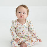 A baby with light hair sits barefoot on a white floor, wearing cream-colored Cactus Kisses Western Print Elastic Waist Leggings by Mabel and Honey, adorned with colorful animals and playful western-inspired patterns, gazing slightly upward with a curious expression.