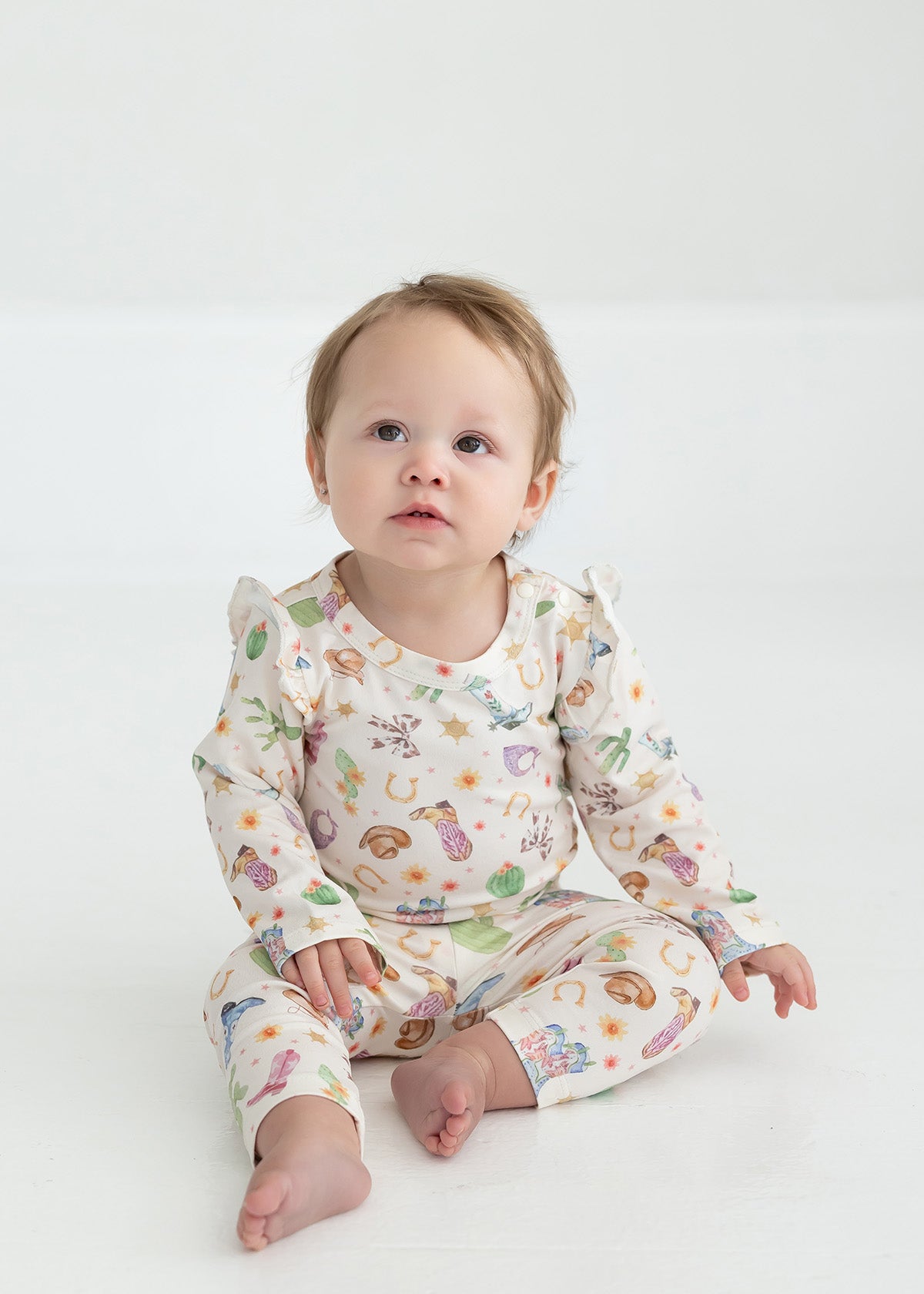 A baby with light hair sits barefoot on a white floor, wearing cream-colored Cactus Kisses Western Print Elastic Waist Leggings by Mabel and Honey, adorned with colorful animals and playful western-inspired patterns, gazing slightly upward with a curious expression.