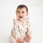 A baby sits on a white floor, smiling and looking slightly upward, dressed in the Mabel and Honey Cactus Kisses Long Sleeve Print Ruffle Neck Bubble Romper.
